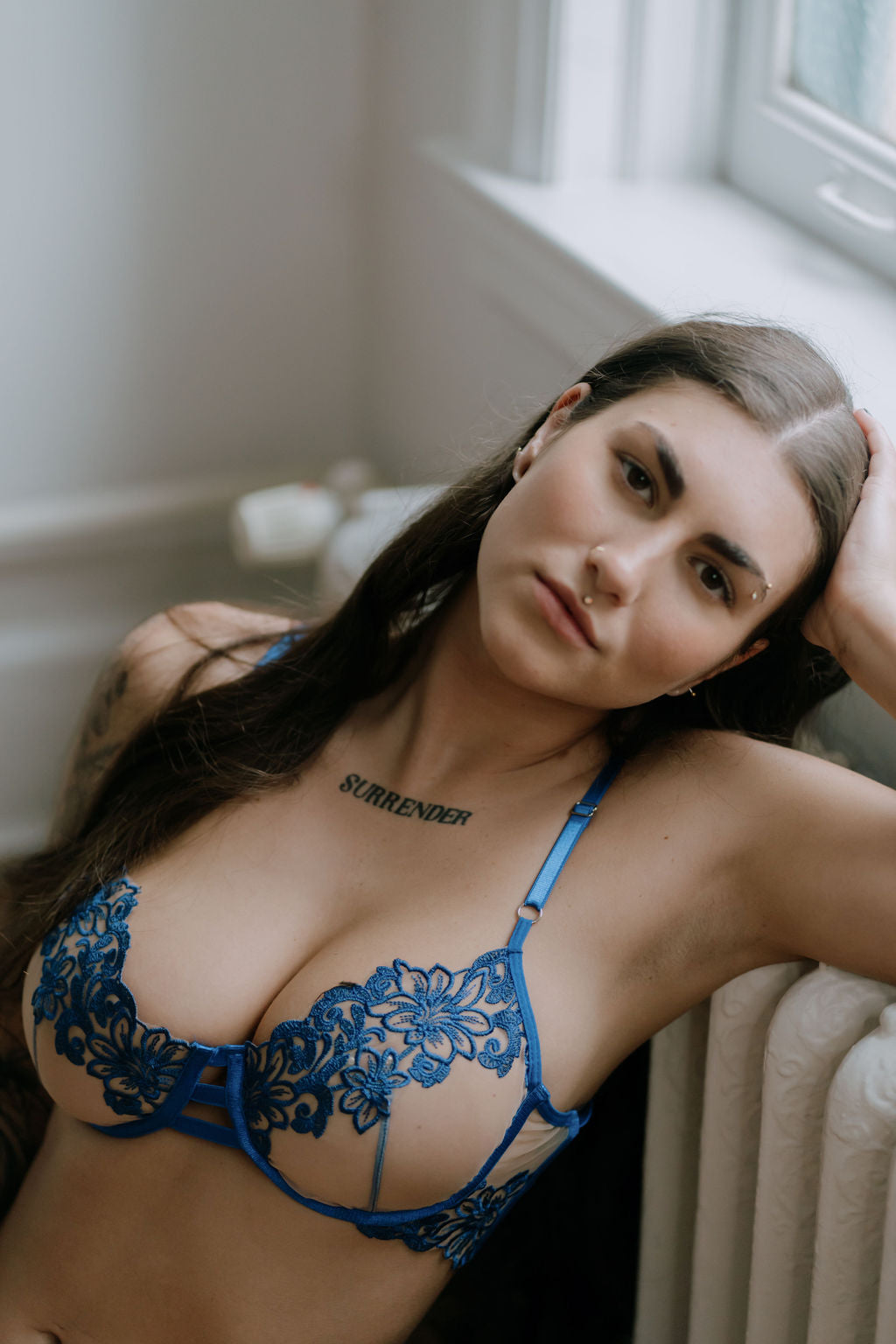 Woman in blue lace lingerie leaning against a radiator in a sunlit room.